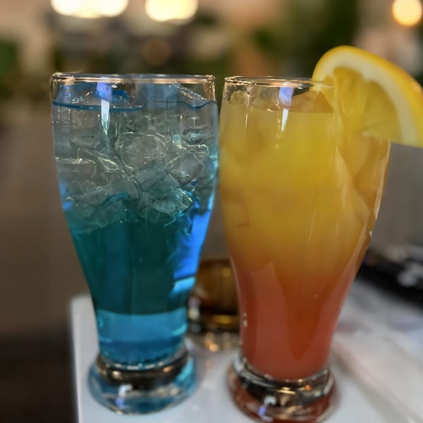 Two vibrant cocktails, one blue and one orange with a lemon slice, on a white surface.
