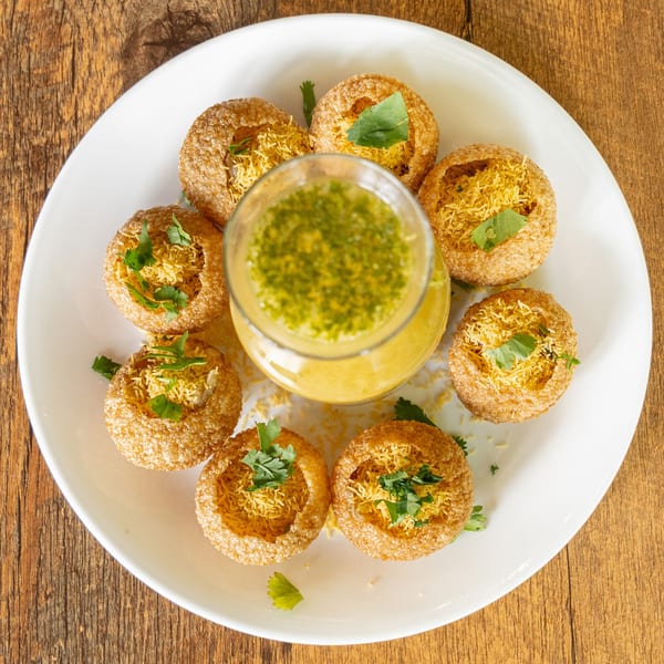 Top-down view of Kinara's Pani Puri, golden crispy shells filled with sev and cilantro, surrounding a glass of green pani.