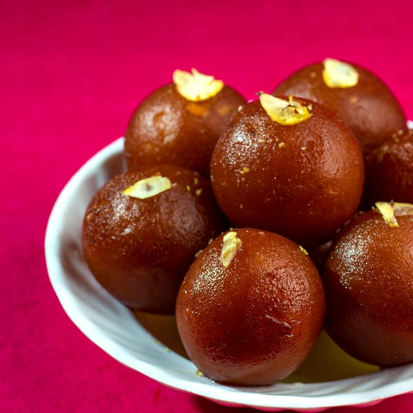 A white bowl filled with glistening brown gulab jamun, garnished with slivered nuts, on a pink background.